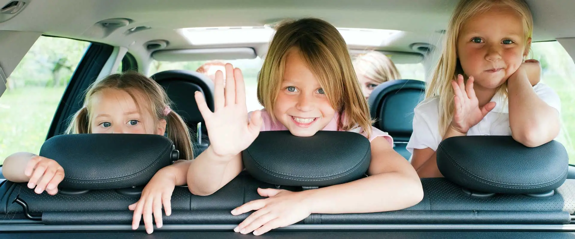 three young girls waving hello from the back seat of a car